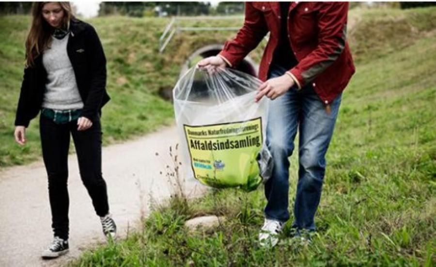 To personer går på en sti med en stor affaldspose med teksten "Affaldsindsamling" på