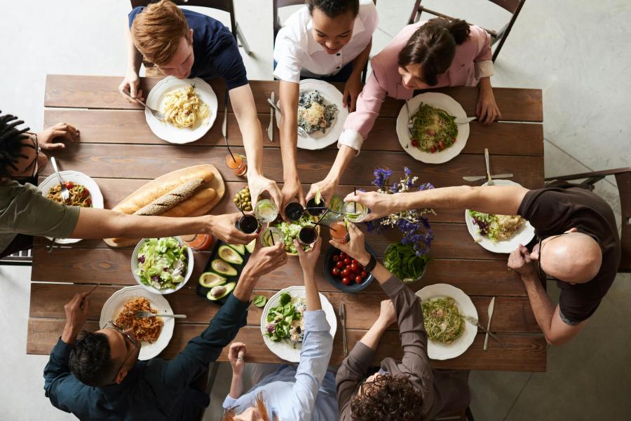 Otte personer set oppefra, der sidder omkring et bord fyldt med mad. De skåler med hinanden og smiler. 