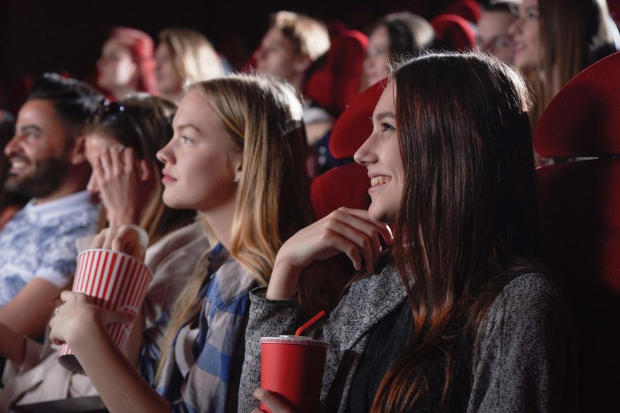 En gruppe mennesker sidder i en biograf. I forgrunden sidder en ung kvinde med en sodavand i hånden. Hun smiler.