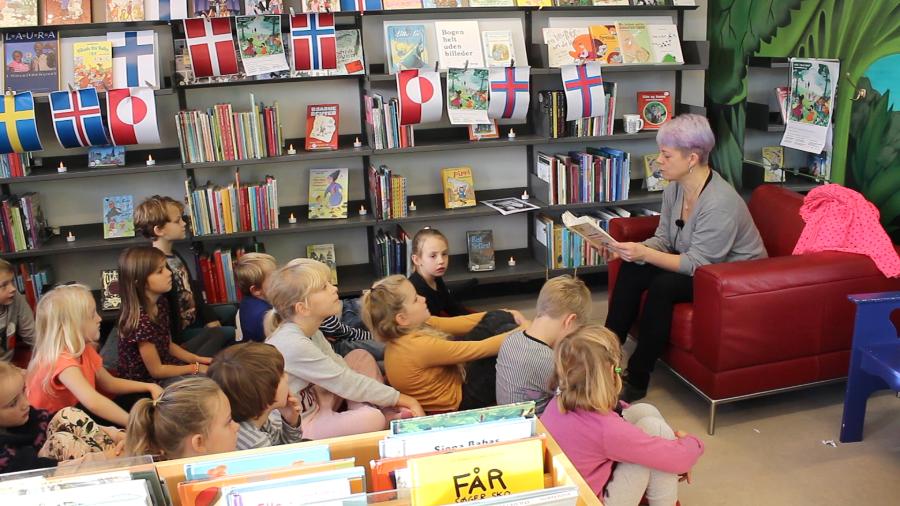 En person læser højt fra en bog for en flok børn som sidder i et bibliotek. Bogreolerne er draperet med flag fra alle de nordiske lande.