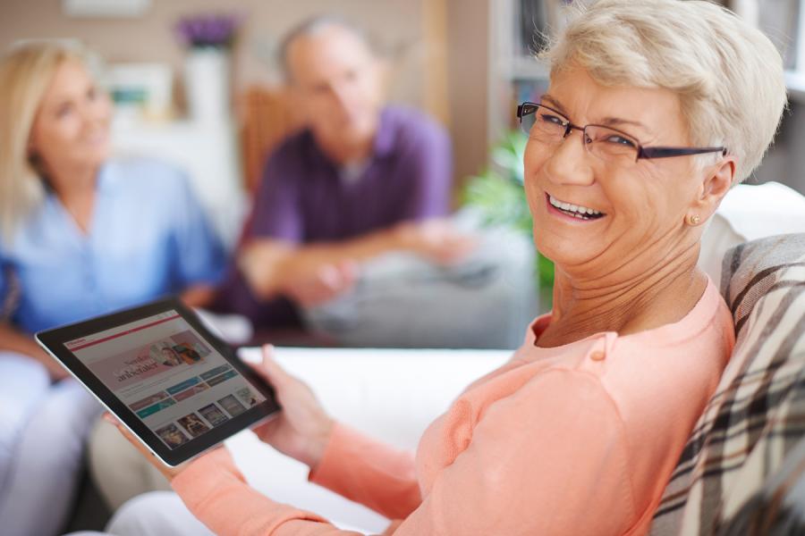 Kvinde med kort lyst hår og briller smiler til kameraet, mens hun holder på en iPad. I baggrunden ses 2 uskarpe personer