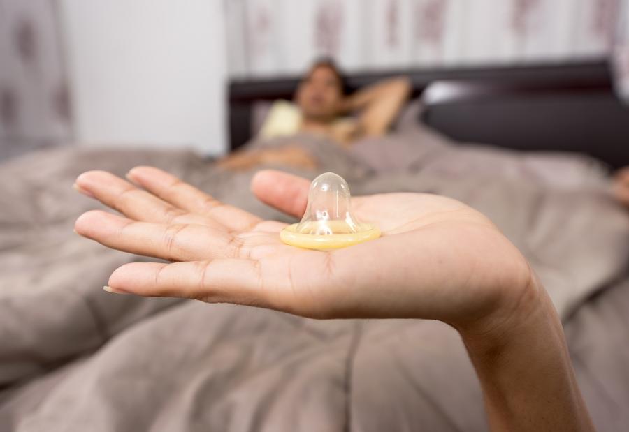 I forgrunden ses en håndflade hvorpå, der ligger et kondom. I baggrunden, ude af fokus, ligger en mand med bar overkrop i en seng.