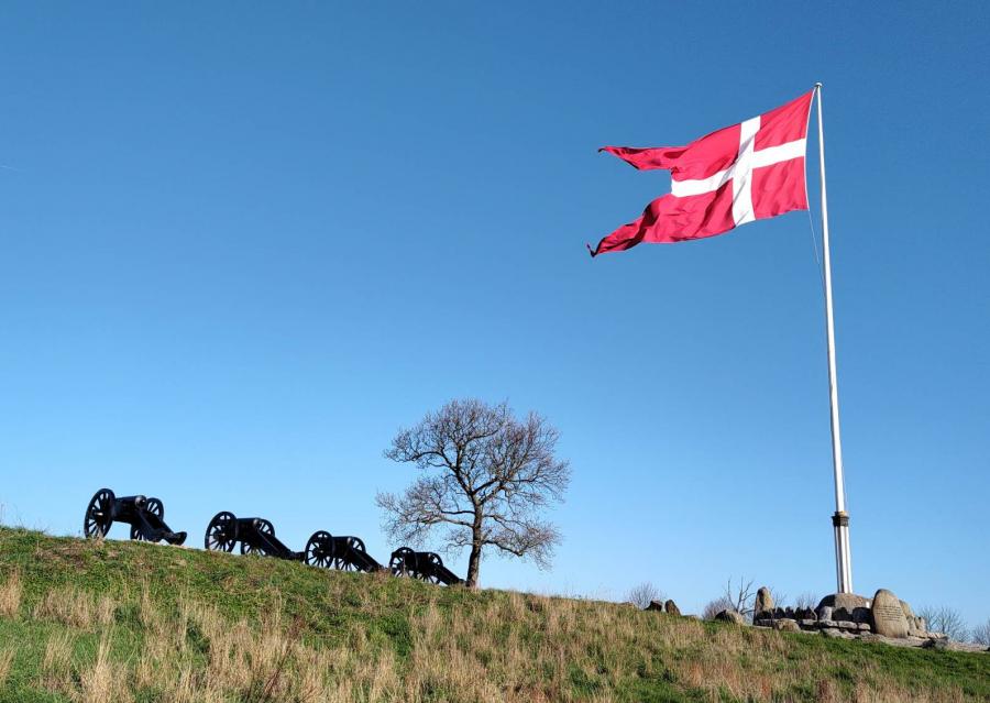 Dronningeflaget vajer på Dybbøl Banke 18. april 2023. I silhouet ses 4 kanoner.
