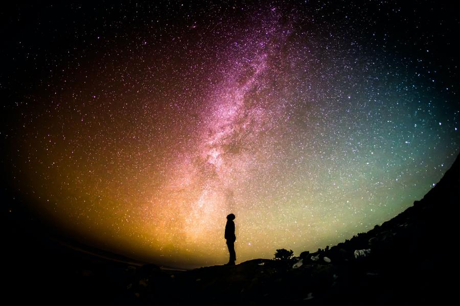 Et menneske står i silhouet og kigger op på en farverig stjernehimmel.