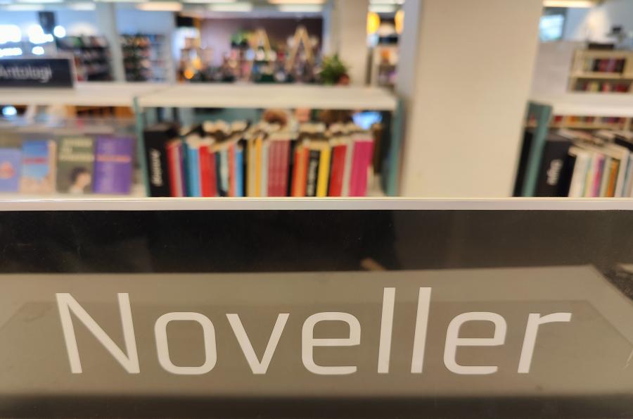 Skilt med teksten "Noveller". I baggrunden anes bibliotekets hylder med bøger,