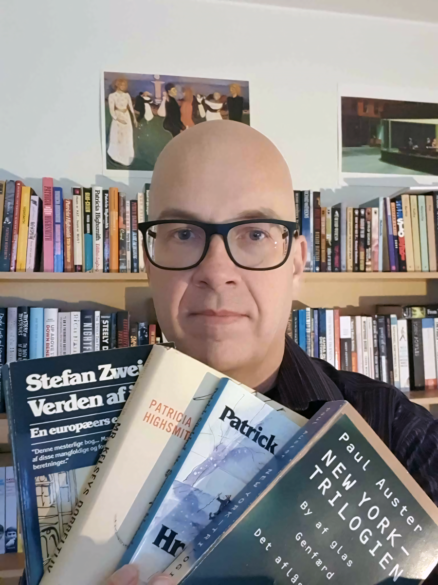 Peter Wollerup står foran en bogreol og holder fire bøger op foran sig. Den forreste bog kan man se er "New York-trilogien" af Pau Auster