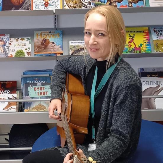 Rikke sidder med en guitar i hænderne. I baggrunden ses en bogreol.