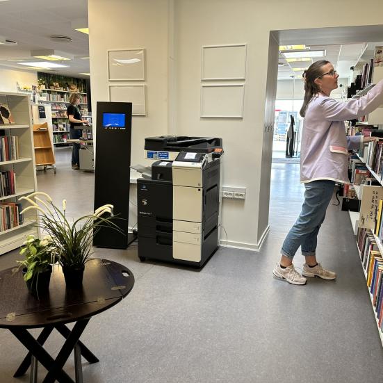 Printeren på Svenstrup Bibliotek står i midten af billedet. I højre side står en kvinde og rækker ud efter en bog på hylden.