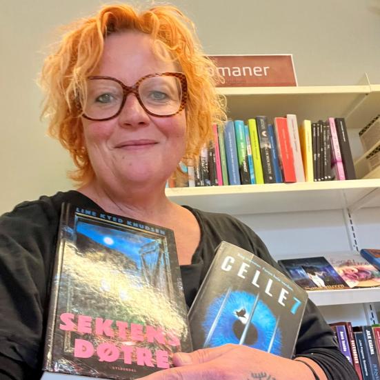 Maria holder bøgerne "Sektens døtre" og "Celle 7" i favnen. Hun står foran en reol med et rødt skilt med teksten "Romaner".