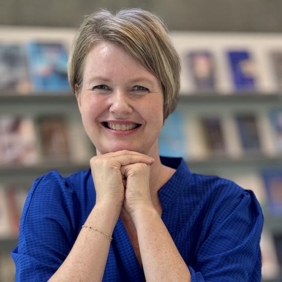 Billede af Dorthe Iversen. Hun hviler albuerne på et bord, hænderne er foldet under hagen. Hun har en blå skjorte på. Hun smiler. 