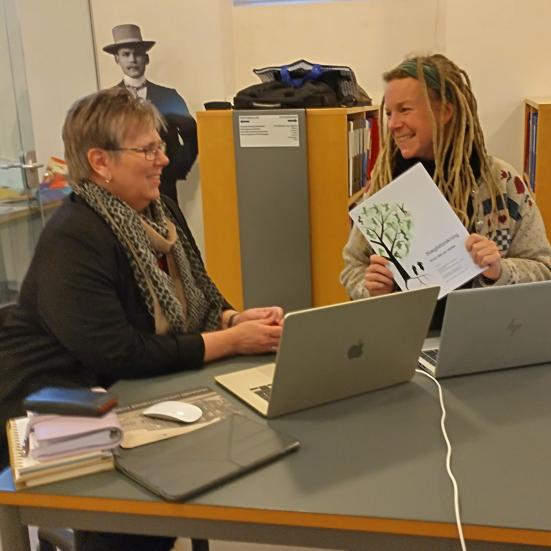 En bibliotekar hjælper en person med slægtsforskning på HistorieAalborg. De sidder begge ved et bord med computere og bøger foran sig. De smiler til hinanden.