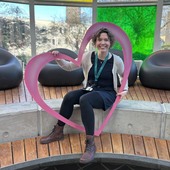 Ditte sidder i glashjørnet på Hovedbiblioteket. Hun holder et stort pink hjerte og smiler. Bag hende ses glashjørnets farverige vinduer.