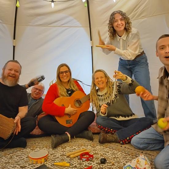En gruppe på seks mennesker sidder og står i et tælt og spiller på hvert sit instrument. Trommer, rasleæg, triangel og guitar