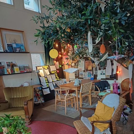 Forstuen i Folkestuen. En lænestol, stuebord og stole, en palme, lamper og andre hyggelige genbrugsmøbler.