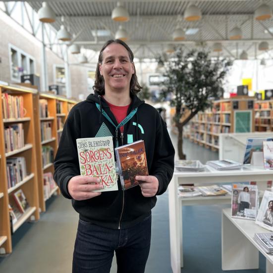 Kenneth står på Hovedbiblioteket. Han holder to af Jens Blendstrups romaner foran sig og smiler til kameraet. Bag Kenneth ses biblioteksrummet med fyldte bogreoler og bogudstillinger.