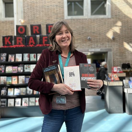 Hanne står foran Ordkraft-udstillingen på Hovedbiblioteket. I sin favn holder hun alle tre romaner, som er omtalt i denne artikel. Hanna smiler og kigger til højre for fotografen. Baggrunden er let sløret og viser udover udstillingen biblioteksrummet.