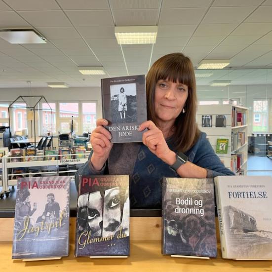 Karin står på biblioteket med et udvalg af Pia Grandjeans bøger stående foran sig med forsiderne frem. Hun holder "Den ariske jøde" op. Bag Karin ses biblioteksrummet.