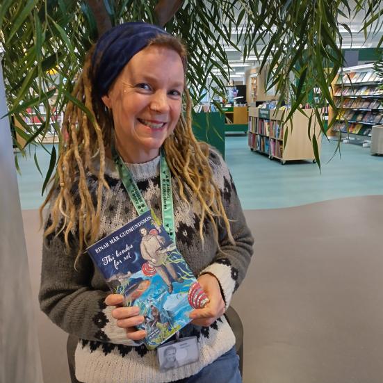 Karina står i afdelingen for skønlitteratur for voksne på Hovedbiblioteket. Hun holder "Thi kendes for ret" i hænderne og smiler til kameraet.