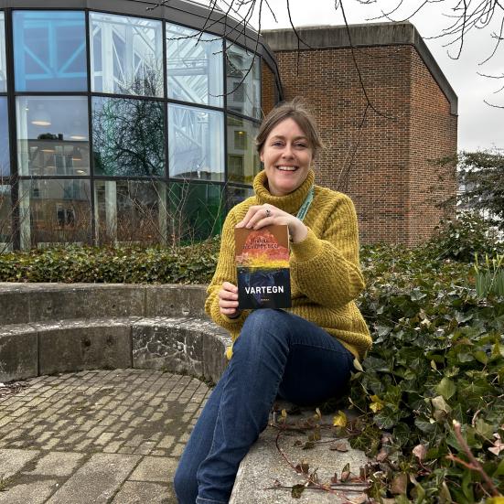 Line sidder i bibliotekshaven ved Hovedbiblioteket. Hun smiler og holder "Vartegn" mellem hænderne med forsiden frem. Bag Line ses Glashjørnets farverige vinduer. 