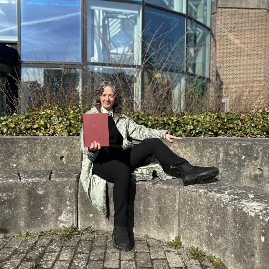 Majken sidder i bibliotekshaven ved Hovebiblioteket. Hun holder bogen "Bror" frem for sig og smiler. Bag Majken ses glashjørnets farverige vinduer. Himlen er blå.