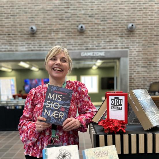 Marianne står ved Ordkraft-udstillingen på Hovedbiblioteket, hvor der er udstillet bøger af forfattere, der gæster festivalen i går. Marianne smiler og holder "Missionær" i hænderne. Bag hende ses biblioteksrummet med Gamezone, som er let sløret.