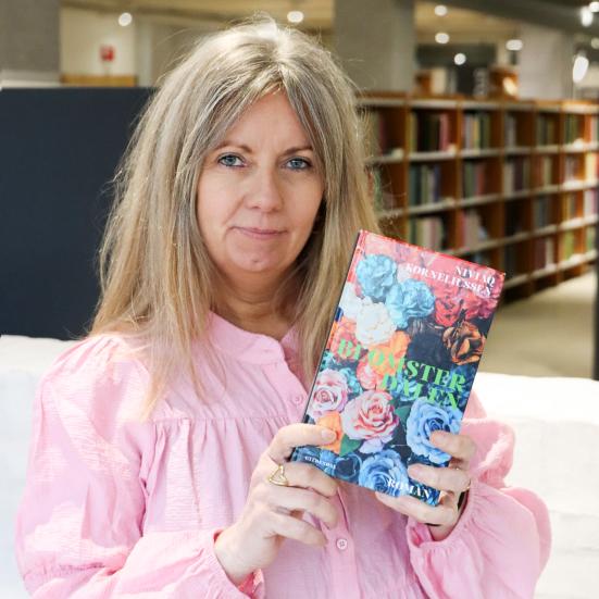 Merete holder bogen "Blomsterdalen" op ved siden af sit hoved, så bogforsiden ses tydeligt. Bag Merete ses biblioteksrummet med fylde bogreoler. Merete kigger direkte ind i kameraet. 