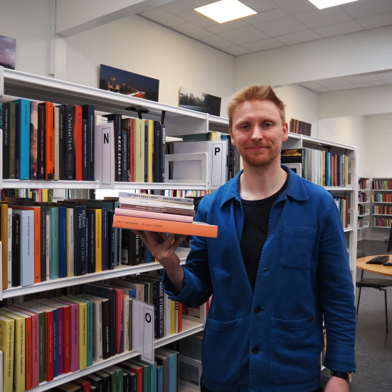 Patrick står med en stak af Dorthe Nors bøger. Bag ham ses biblioteksrummet med fyldte bogreoler. Han kigger ind i kameraet og smiler.