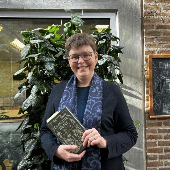 Vita står foran de grønne planter foran Makerspace på Hovedbiblioteket. Hun smiler og holder bogen "Ulvemælk" i hænderne. Lyset falder flot på væggen bag hende.