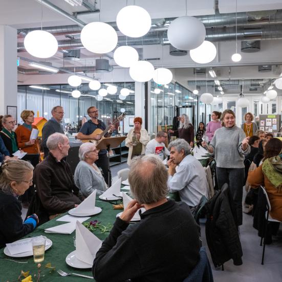 Foto af mange forskellige mennesker, der mødes til fællesspisning og møde. En kvinde holder velkomsttale i midten af forsamlingen og alles opmærksomhed er rettet mod hende.