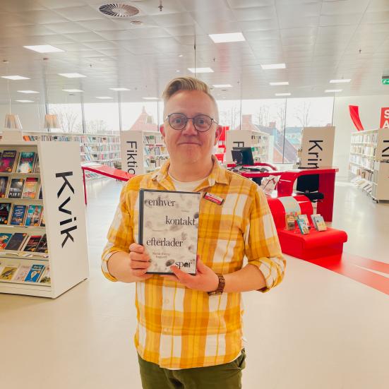 Martin står på Hjørring Bibliotek. Han holder den omtalte bog i denne artikel mellem hænderne. Bag Martin ses biblioteksrummet med fyldte bogreoler og udstillinger.
