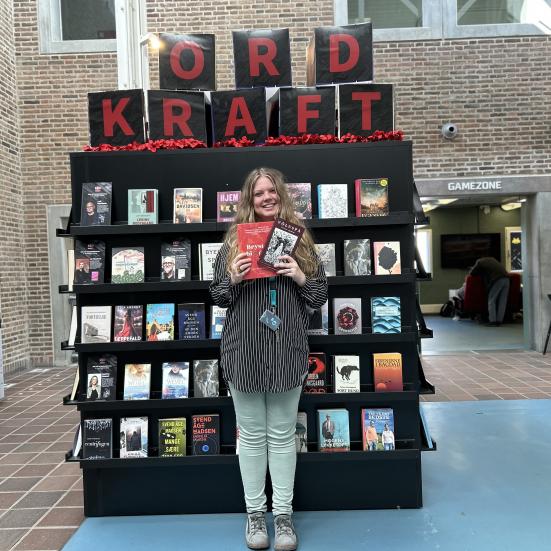 Sørine står foran Ordkraft udstillingen på Hovedbiblioteket. Udstillingen viser et stort udvalg af bøger skrevet af forfattere, der gæster festivalen i år. Sørine holder de to omtalte romaner i denne artikel i hænderne og smiler. Bag hende ses biblioteksrummet med bl.a. Gamezone.