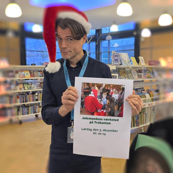 En person, Jes, står på Trekanten Bibliotek iført nissehue og poserende med en plakat. På plakaten står: "Julemandens værksted på Trekanten. Lørdag den 3. december Kl. 10-14"