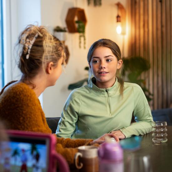 Et barn ig en voksen sidder ved et bord og taler. I forgrunden ses fra ryggen endnu et barn, der sidder med en tablet. 