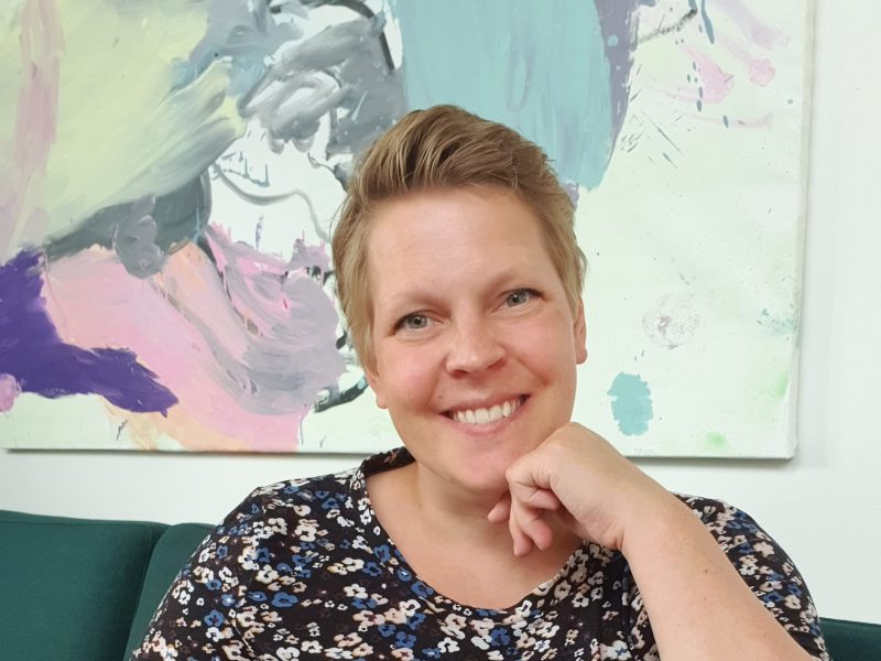 Birgitte sidder i en sofa foran et maleri og smiler.