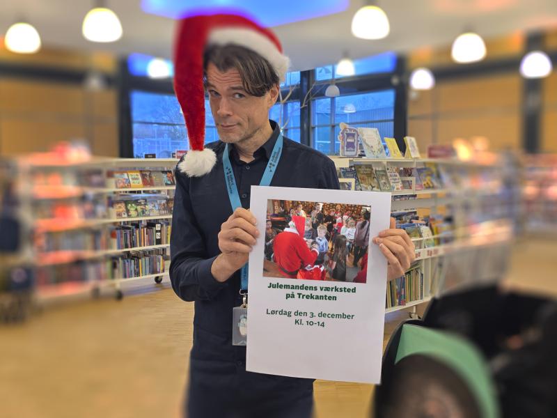 En person, Jes, står på Trekanten Bibliotek iført nissehue og poserende med en plakat. På plakaten står: "Julemandens værksted på Trekanten. Lørdag den 3. december Kl. 10-14"
