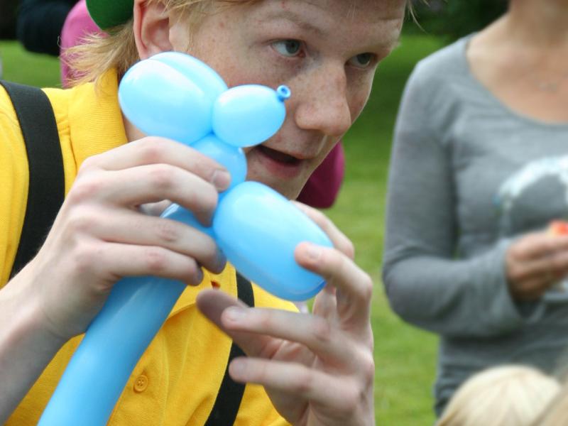 En person iført gul polo, sorte seler og grøn bowlerhat er i færd med at lave et ballondyr. Der er børn og voksne på billedet i baggrunden.
