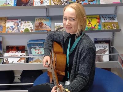 Rikke sidder med en guitar i hænderne. I baggrunden ses en bogreol.