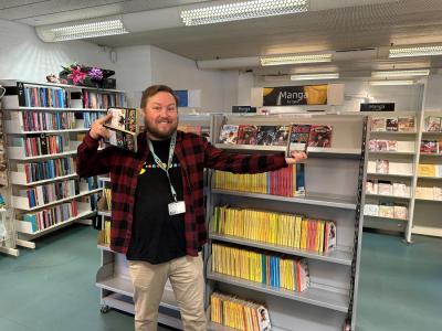 Kristian står smilende med tre mangaer i hænderne. Han står foran en bogreol fyldt med mangaer i Voksen Skønlitteratur på Hovedbiblioteket.
