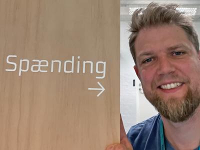 En mand står ved siden af en bogreol med teksten "spænding" og en pil, der peger mod mandens hoved.