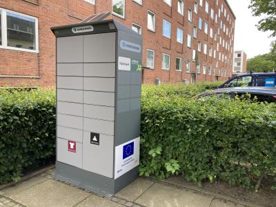 En pakkeboks står ved en parkeringsplads foran en etagebygning.