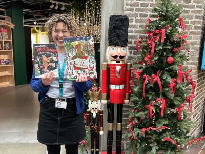 Ditte står foran indgangen til børnebiblioteket på Hovedbiblioteket. Ved siden af hende ses juletræ og julepynt. Hun holder bøgerne "Iqbal Farooq og julesvineriet" og "Hvor er julepøllerne?" frem for sig, mens hun smiler.