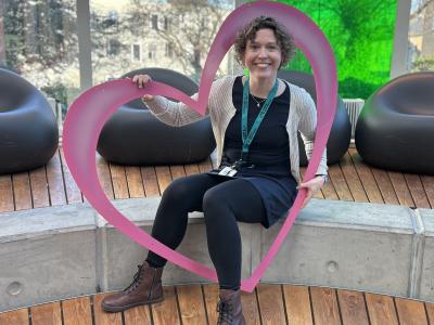 Ditte sidder i glashjørnet på Hovedbiblioteket. Hun holder et stort pink hjerte og smiler. Bag hende ses glashjørnets farverige vinduer.