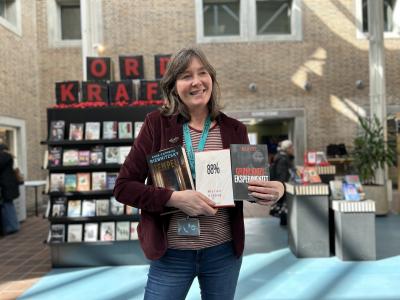 Hanne står foran Ordkraft-udstillingen på Hovedbiblioteket. I sin favn holder hun alle tre romaner, som er omtalt i denne artikel. Hanna smiler og kigger til højre for fotografen. Baggrunden er let sløret og viser udover udstillingen biblioteksrummet.