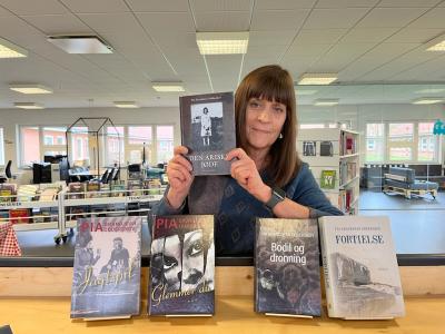 Karin står på biblioteket med et udvalg af Pia Grandjeans bøger stående foran sig med forsiderne frem. Hun holder "Den ariske jøde" op. Bag Karin ses biblioteksrummet.
