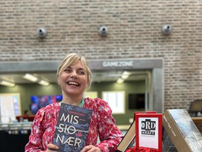 Marianne står ved Ordkraft-udstillingen på Hovedbiblioteket, hvor der er udstillet bøger af forfattere, der gæster festivalen i går. Marianne smiler og holder "Missionær" i hænderne. Bag hende ses biblioteksrummet med Gamezone, som er let sløret.