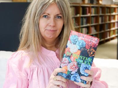 Merete holder bogen "Blomsterdalen" op ved siden af sit hoved, så bogforsiden ses tydeligt. Bag Merete ses biblioteksrummet med fylde bogreoler. Merete kigger direkte ind i kameraet. 