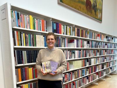 Michelle står på biblioteket. Hun holder "Øvelser i mørke" og "Poesibog" i hænderne og kigger smilende ind i kameraet. Bag Michelle ses fyldte bogreoler og et stort maleri på væggen.