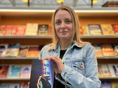 Nina står foran en bogreol på Hovedbiblioteket. Hun holder bogen "Esbens fire liv" i hænderne og smiler. Baggrunden er let sløret.