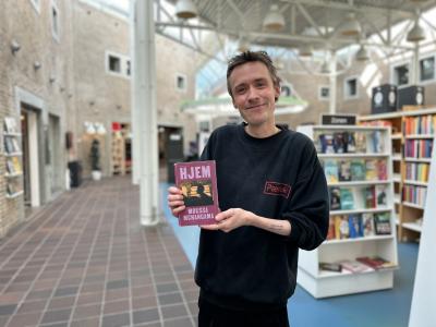 Søren Malthe står med den omtalte bog i denne artikel i hænderne. Han smiler til kameraet. Baggrunden er let sløret og viser biblioteksrummet.