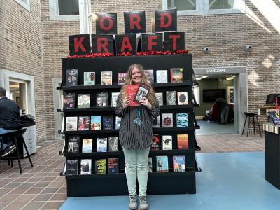 Sørine står foran Ordkraft udstillingen på Hovedbiblioteket. Udstillingen viser et stort udvalg af bøger skrevet af forfattere, der gæster festivalen i år. Sørine holder de to omtalte romaner i denne artikel i hænderne og smiler. Bag hende ses biblioteksrummet med bl.a. Gamezone.